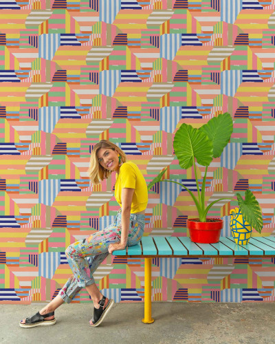 Femeie zâmbitoare așezată pe o bancă albastră în fața unui perete acoperit cu tapet cu dungi și forme geometrice colorate.