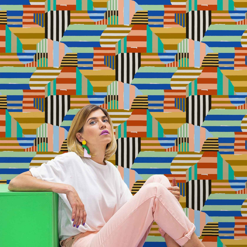 Femeie zâmbitoare așezată pe o bancă albastră în fața unui perete acoperit cu tapet cu dungi și forme geometrice colorate.