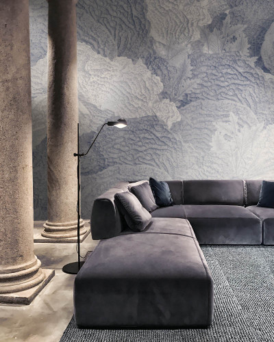 Tapet contemporan cu design abstract, care prezintă frunze în nuanțe delicate de albastru și gri. Tapetul este aplicat pe un perete din spatele unui canapea gri în formă de L.