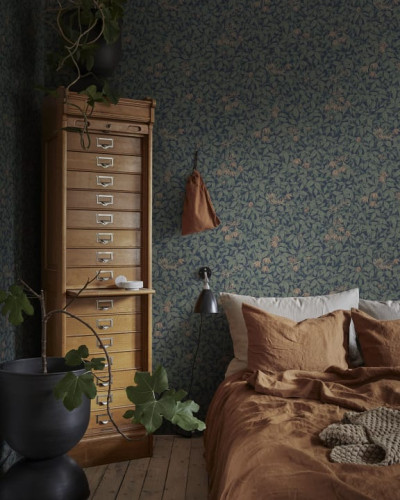 Tapet Linnea Midnight Blue cu model dens de frunze, flori și plante mici, într-o nuanță albastră închisă, decorând un dormitor cu patul aranjat cu lenjerie de pat maro.