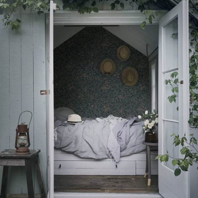 Tapet Linnea Midnight Blue cu model dens de frunze, flori și plante mici, într-o nuanță albastră închisă, decorând un dormitor cu patul aranjat cu lenjerie de pat maro.