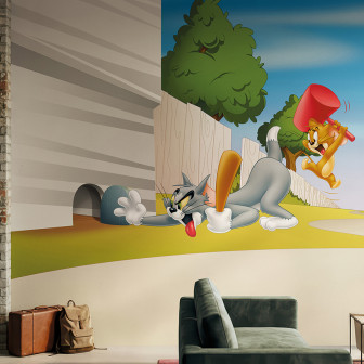Fototapet vibrant cu Tom, pisica gri cu bâtă de baseball, și Jerry, șoricelul portocaliu cu ciocan roșu, într-o scenă din desene animate. Decor modern într-un spațiu de birou sau living cu un perete cărămidă aparentă și canapea verde.