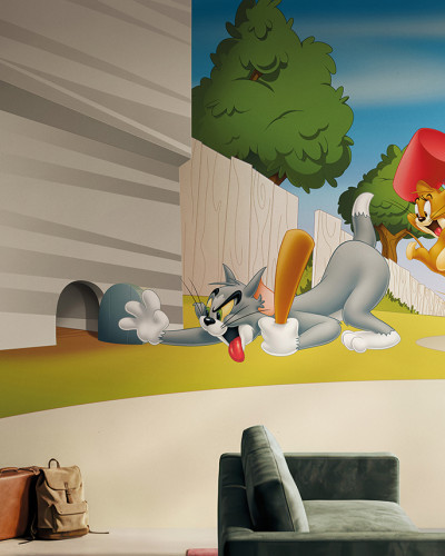 Fototapet vibrant cu Tom, pisica gri cu bâtă de baseball, și Jerry, șoricelul portocaliu cu ciocan roșu, într-o scenă din desene animate. Decor modern într-un spațiu de birou sau living cu un perete cărămidă aparentă și canapea verde.