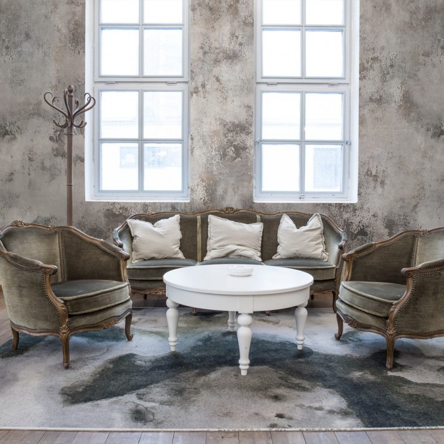 Tapet 3D Patina, cu aspect de beton, într-un dormitor cu pat dublu, cu lenjerie de pat albastră, perne decorative și o canapea cu o pătură de blană