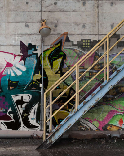 Tapet foto cu design urban - Stairway Graffiti, care prezintă un perete cu grafitti colorat, o scară metalică și beton brut.