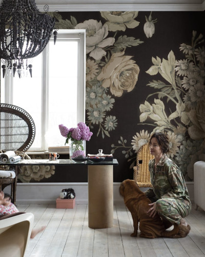 Imagine cu un tapet floral cu flori mari și detalii elegante, în culori intense pe fundal închis. Tapetul este lipit pe un perete dintr-o cameră, care include o canapea, o masă și o lampă.
