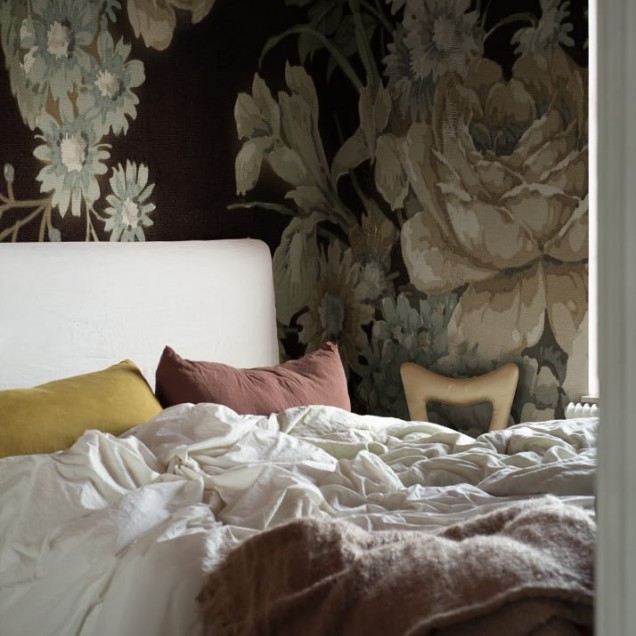 Imagine cu un tapet floral cu flori mari și detalii elegante, în culori intense pe fundal închis. Tapetul este lipit pe un perete dintr-o cameră, care include o canapea, o masă și o lampă.