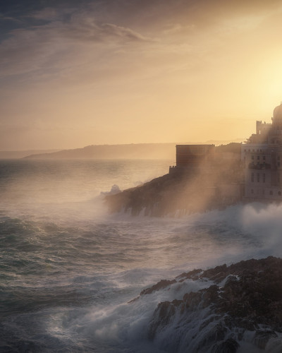 Tapet cu imagine a unei linii de coastă dramatice, cu ape tulburi, un oraș fermecător în depărtare și razele soarelui strălucind prin nori.