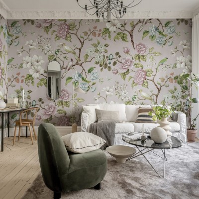 Tapet floral Grandiflora, roz pal, cu flori roz și albe, păsări și frunze verzi, decorând un living cu o canapea alba și un fotoliu verde.