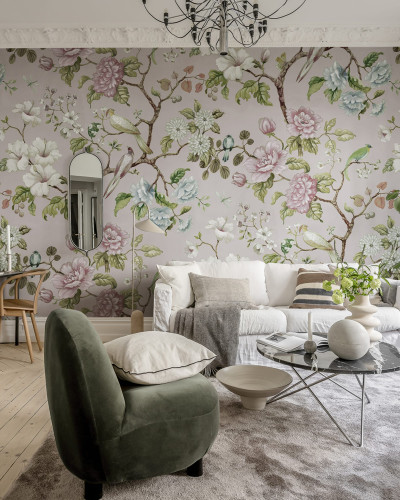 Tapet floral Grandiflora, roz pal, cu flori roz și albe, păsări și frunze verzi, decorând un living cu o canapea alba și un fotoliu verde.
