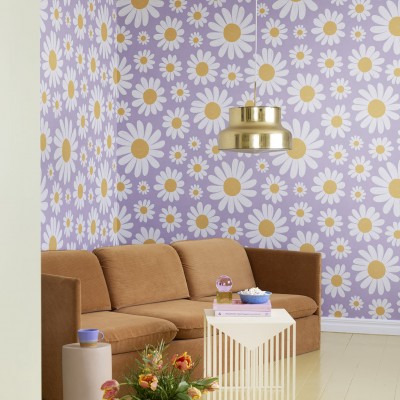 Tapet floral cu margarete albe pe fond liliac, decorand un living cu canapea maro si o masa cu flori.