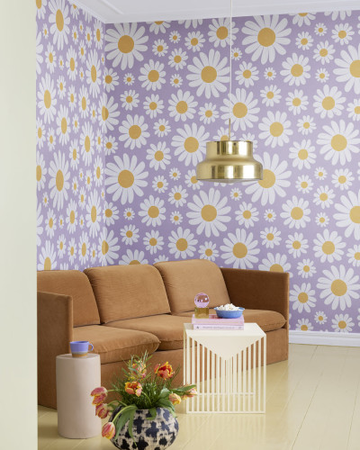 Tapet floral cu margarete albe pe fond liliac, decorand un living cu canapea maro si o masa cu flori.