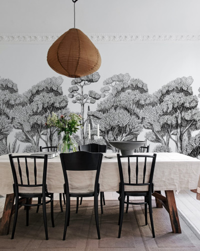 Tapet Forest Park Graphite cu un design minimalist ce prezintă copaci în tonuri de gri. Tapetul este aplicat pe un perete într-o sufragerie modernă cu o masă din lemn, scaune negre și o lampă cu abajur din hârtie.