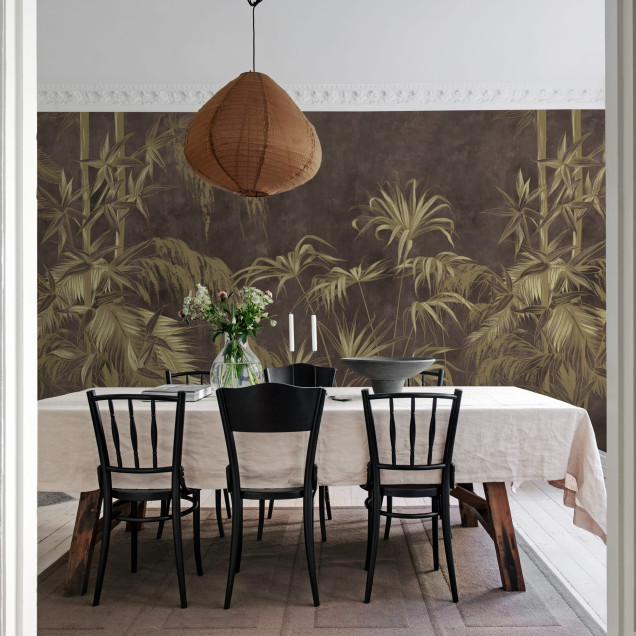 Un tapet cu un design exotic, Golden Jungle, Dark, care prezintă frunze aurii pe un fundal brun-cenusiu. Tapetul este amplasat pe un perete din living, alături de o canapea albă, o masă de cafea și o bibliotecă.