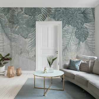 Fototapet Monstera Wall, Blue într-un living modern cu canapea bej, masă de cafea cu blat de marmură și covor albastru. Peretele accent este decorat cu frunze mari de monstera și palmier pe un fundal texturat, gri-albăstrui.