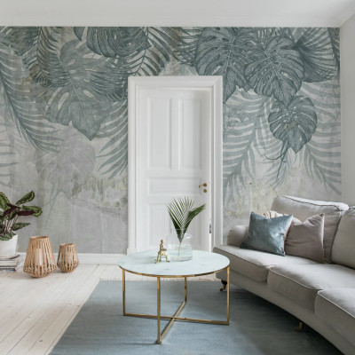 Fototapet Monstera Wall, Blue într-un living modern cu canapea bej, masă de cafea cu blat de marmură și covor albastru. Peretele accent este decorat cu frunze mari de monstera și palmier pe un fundal texturat, gri-albăstrui.