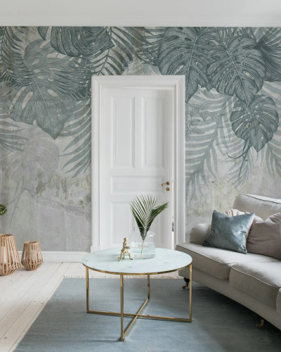 Fototapet Monstera Wall, Blue într-un living modern cu canapea bej, masă de cafea cu blat de marmură și covor albastru. Peretele accent este decorat cu frunze mari de monstera și palmier pe un fundal texturat, gri-albăstrui.