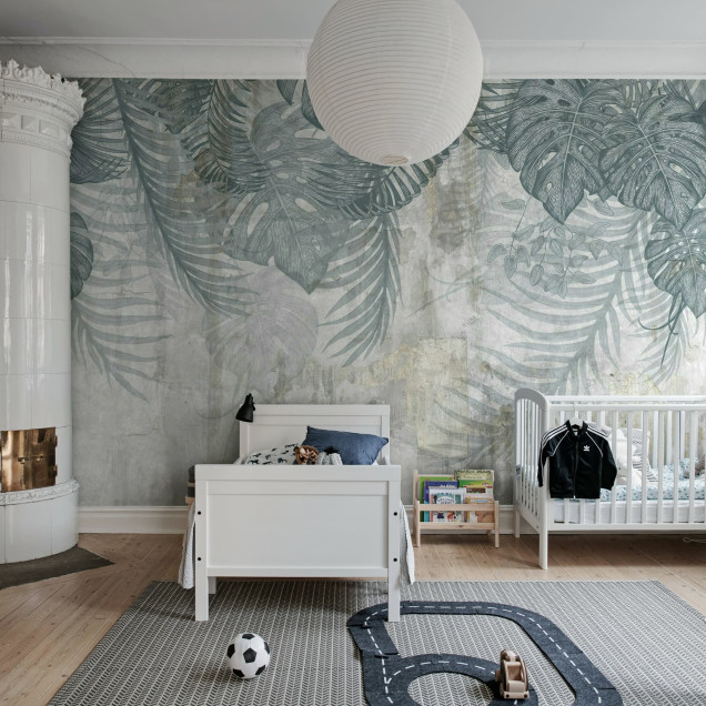 Fototapet Monstera Wall, Blue într-un living modern cu canapea bej, masă de cafea cu blat de marmură și covor albastru. Peretele accent este decorat cu frunze mari de monstera și palmier pe un fundal texturat, gri-albăstrui.