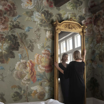Femeie privindu-se în oglindă aurie într-o cameră decorată cu tapet floral Ines, Sage Green, cu motive ample pe fond verde salvie și grinzi de lemn expuse.