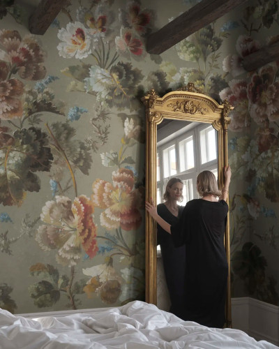 Femeie privindu-se în oglindă aurie într-o cameră decorată cu tapet floral Ines, Sage Green, cu motive ample pe fond verde salvie și grinzi de lemn expuse.