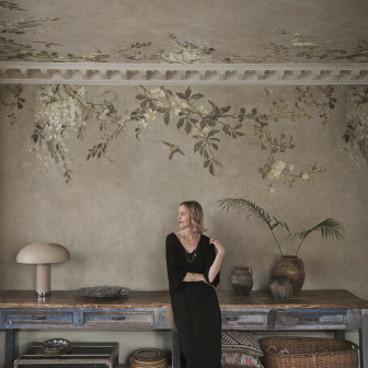 Cameră decorată cu tapet Luca Square, Beige, cu model floral și păsări pe pereți și tavan. O femeie elegantă se sprijină de o masă rustică. Design interior sofisticat, natură, culori pământii.