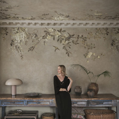 Cameră decorată cu tapet Luca Square, Beige, cu model floral și păsări pe pereți și tavan. O femeie elegantă se sprijină de o masă rustică. Design interior sofisticat, natură, culori pământii.