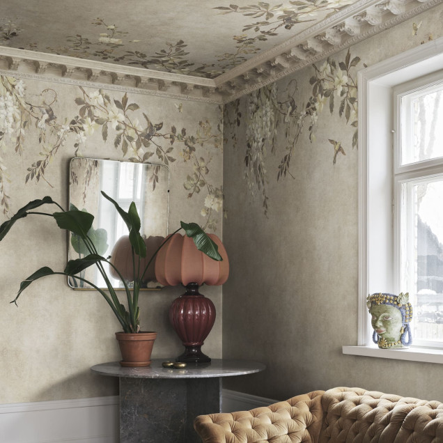 Cameră decorată cu tapet Luca Square, Beige, cu model floral și păsări pe pereți și tavan. O femeie elegantă se sprijină de o masă rustică. Design interior sofisticat, natură, culori pământii.