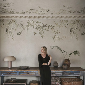 Femeie blondă într-un living elegant cu pereți și tavan acoperiți de tapet Luca Square, Sand, prezentând un design floral cu ramuri, flori delicate și păsări pe un fundal bej nisipiu, alături de un bufet din lemn vechi și obiecte de decor vintage.