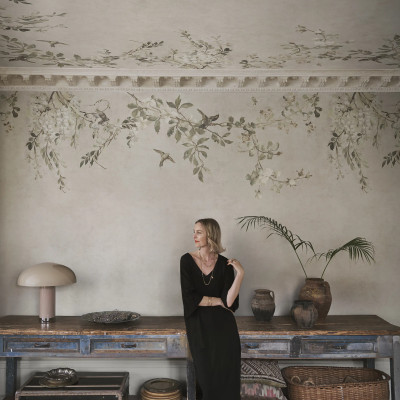 Femeie blondă într-un living elegant cu pereți și tavan acoperiți de tapet Luca Square, Sand, prezentând un design floral cu ramuri, flori delicate și păsări pe un fundal bej nisipiu, alături de un bufet din lemn vechi și obiecte de decor vintage.