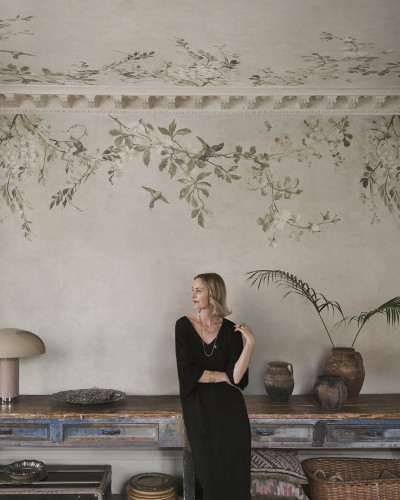 Femeie blondă într-un living elegant cu pereți și tavan acoperiți de tapet Luca Square, Sand, prezentând un design floral cu ramuri, flori delicate și păsări pe un fundal bej nisipiu, alături de un bufet din lemn vechi și obiecte de decor vintage.