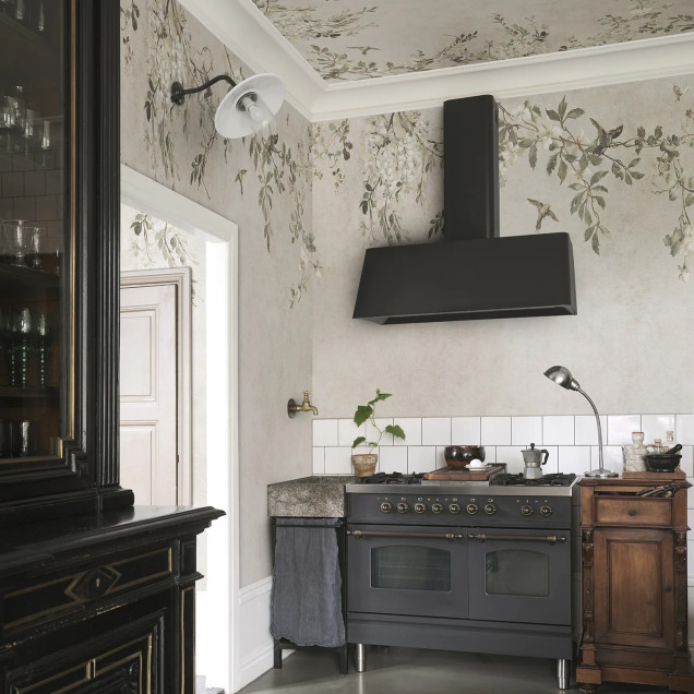 Femeie blondă într-un living elegant cu pereți și tavan acoperiți de tapet Luca Square, Sand, prezentând un design floral cu ramuri, flori delicate și păsări pe un fundal bej nisipiu, alături de un bufet din lemn vechi și obiecte de decor vintage.
