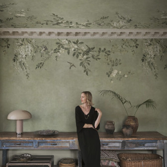 Femeie într-un living elegant cu tapet Luca Rectangle, Sage Green. Peretele și tavanul sunt decorate cu un model botanic de flori albe și păsări pe fundal verde salvie, alături de o consolă vintage și o lampă de masă.