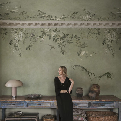Femeie într-un living elegant cu tapet Luca Rectangle, Sage Green. Peretele și tavanul sunt decorate cu un model botanic de flori albe și păsări pe fundal verde salvie, alături de o consolă vintage și o lampă de masă.