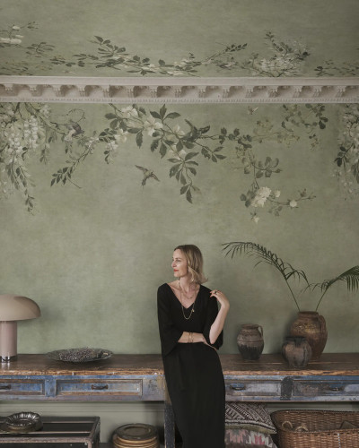 Femeie într-un living elegant cu tapet Luca Rectangle, Sage Green. Peretele și tavanul sunt decorate cu un model botanic de flori albe și păsări pe fundal verde salvie, alături de o consolă vintage și o lampă de masă.