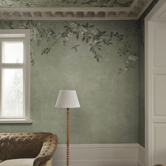 Femeie într-o cameră decorată cu tapet Luca Square Sage Green. Tapetul verde salvie are un model floral delicat cu wisteria și păsări, extins pe tavan, creând un ambient clasic și natural.