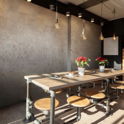 Tapet Marla Moka cu design beton gri deschis, cu cercuri gri închise. Imaginea prezintă un interior modern, cu o masă din lemn și scaune metalice, pe care se află plante verzi.