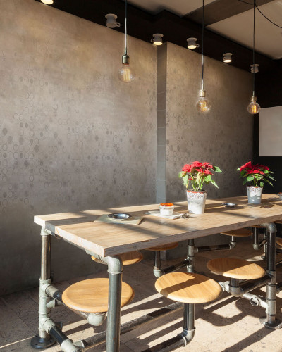 Tapet Marla Moka cu design beton gri deschis, cu cercuri gri închise. Imaginea prezintă un interior modern, cu o masă din lemn și scaune metalice, pe care se află plante verzi.